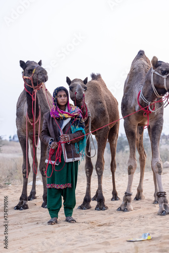 Portrait of an young Indian rajasthani woman in colorful traditional dress carrying camel at Pushkar Camel Fair ground during winter morning.