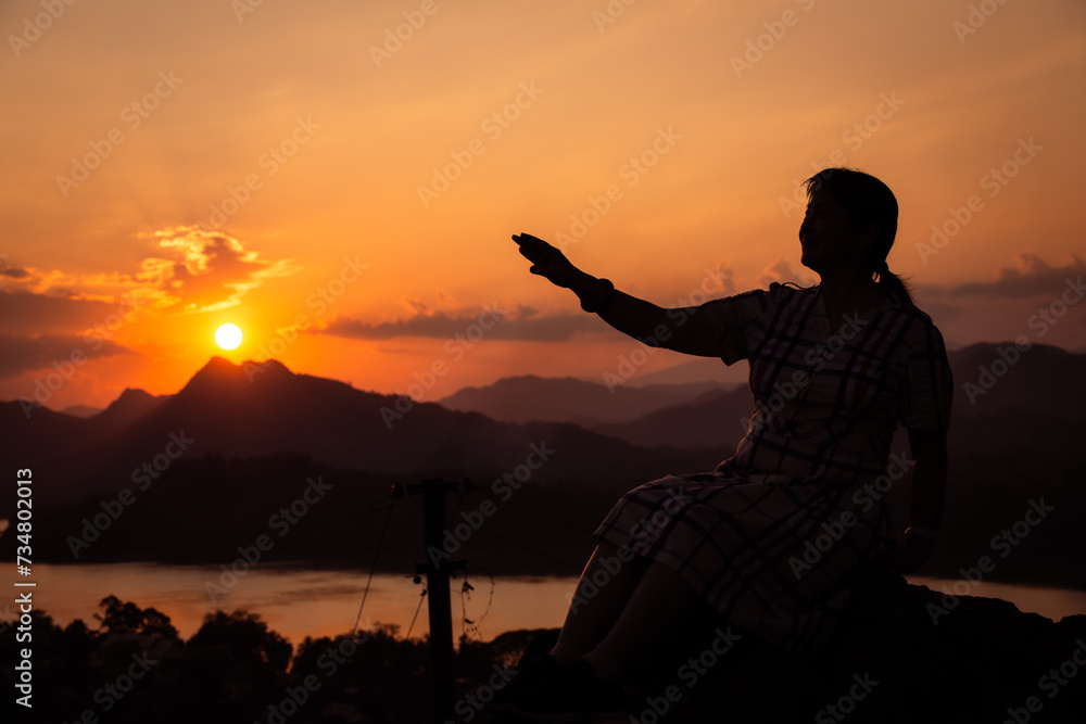 Ombre d'une femme asiatique assise tendant la main vers le coucher de soleil au-dessus d'une montagne