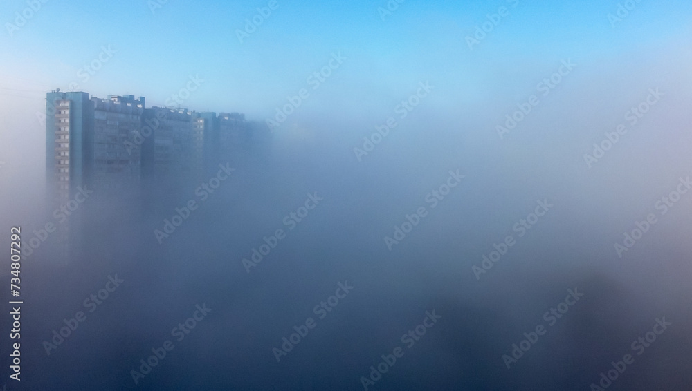 Fototapeta premium A white-and-blue multi-storey residential building is shrouded in fog in the early spring morning.