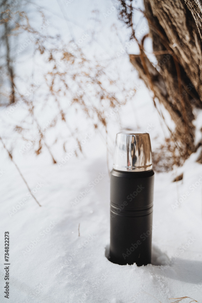 Black thermos bottle with a silver lid sits in the snow near a tree trunk on a sunny winter day. Black thermos bottle with a silver lid sits in the snow near a tree trunk on a sunny winter day.