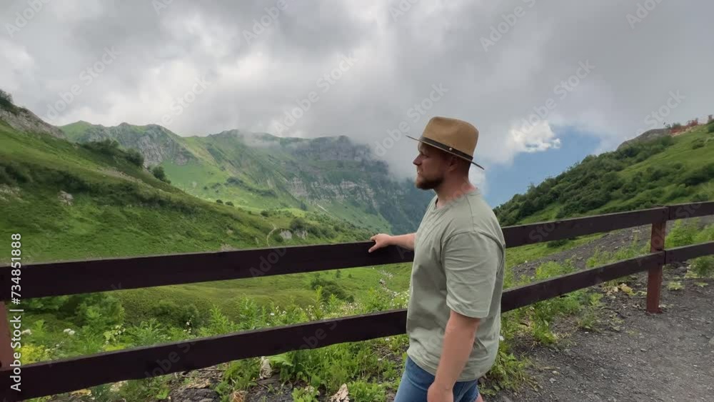 A middle-aged man, a traveler, with a beard, wearing a straw hat walks along a wooden fence in the mountains on a warm summer day in slowmotion A middle-aged man, a traveler, with a beard, wearing a straw hat walks along a wooden fence in the mountains on a warm summer day in slowmotion