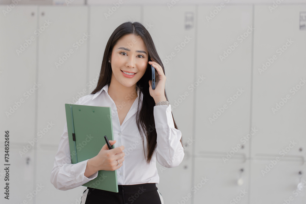 Happy asian young businesswoman holding documents folders in office working space, Asian female employee using laptop talking on the phone at workplace. Happy asian young businesswoman holding documents folders in office working space, Asian female employee using laptop talking on the phone at workplace.