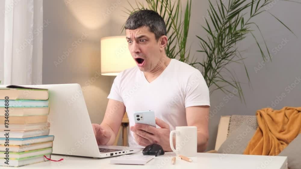 Laptop use for business. Wireless setup at home. Job and lifestyle balance. Caucasian man wearing white t-shirt sitting working on computer and using smartphone at home Laptop use for business. Wireless setup at home. Job and lifestyle balance. Caucasian man wearing white t-shirt sitting working on computer and using smartphone at home