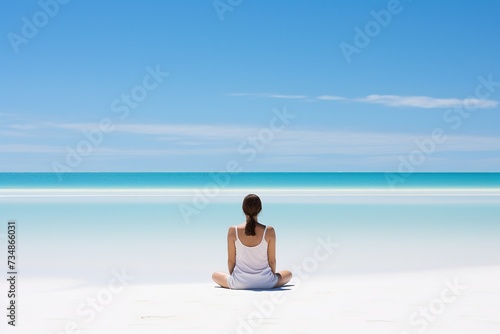 A woman in white summer clothes sits  viewed from the back  in the lotus position on the ocean shore and meditates while looking into the distance. White sandy beach  blue sky with light clouds.