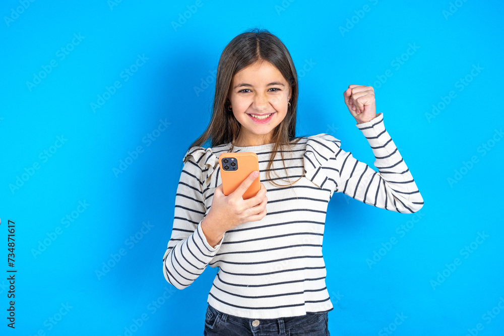 Young beautiful teen girl wearing striped T-shirt holding in hands cell and rising his fist up being excited after reading good news. Young beautiful teen girl wearing striped T-shirt holding in hands cell and rising his fist up being excited after reading good news.