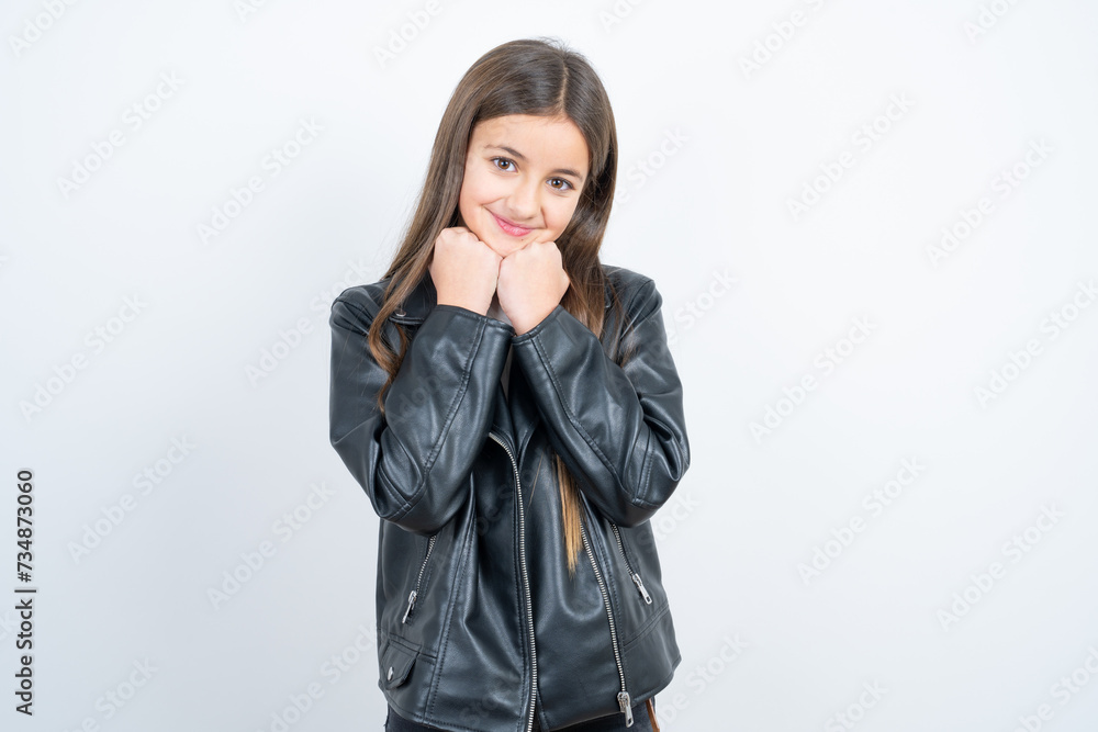 Young beautiful teen girl wearing biker jacket holds hands under chin, glad to hear heartwarming words from stranger Young beautiful teen girl wearing biker jacket holds hands under chin, glad to hear heartwarming words from stranger