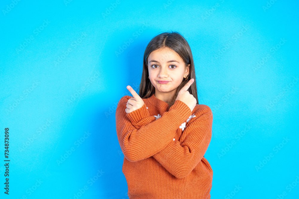 Fototapeta premium Serious Young kid girl wearing orange sweater crosses hands and points at different sides hesitates between two items. Hard decision concept