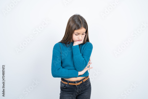 Very bored beautiful kid girl wearing blue sweater holding hand on cheek while support it with another crossed hand, looking tired and sick,