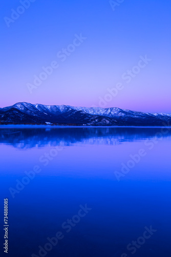 淡い紫色のグラデーションに染まる夜明けの空と雪山を水面に反射する湖。日本の北海道の旅行地である屈斜路湖。雄大で美しい自然。