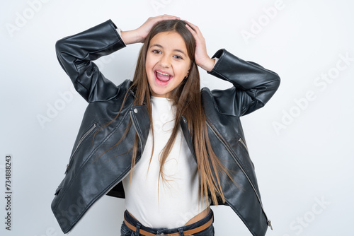 Cheerful overjoyed beautiful kid girl wearing biker jacket reacts rising hands over head after receiving great news.