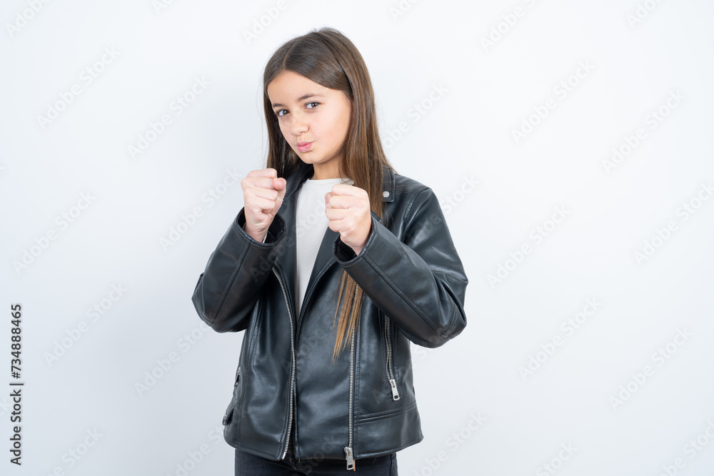 Displeased annoyed beautiful kid girl wearing biker jacket over white background clenches fists, gestures pissed, ready to revenge, looks with aggression at camera stands full of hate, being pressured Displeased annoyed beautiful kid girl wearing biker jacket over white background clenches fists, gestures pissed, ready to revenge, looks with aggression at camera stands full of hate, being pressured