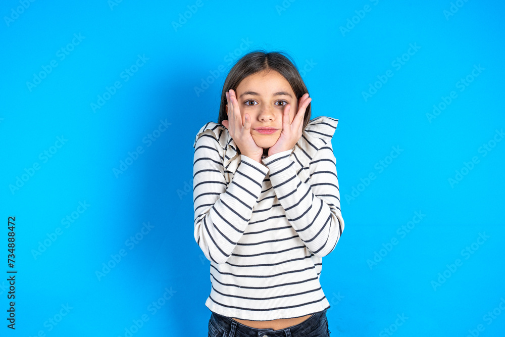 Fototapeta premium Stupefied beautiful kid girl wearing striped T -shirt expresses excitement and thrill, keeps jaw dropped, hands on cheeks, has eyes popped out
