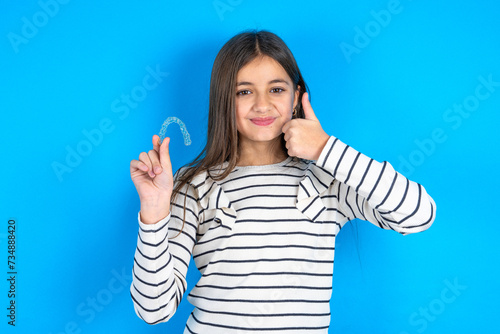 beautiful kid girl wearing  striped T-shirt holding an invisible braces aligner and rising thumb up, recommending this new treatment. Dental healthcare concept.