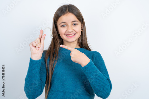 beautiful kid girl wearing blue sweater holding an invisible aligner and pointing at it. Dental healthcare and confidence concept.