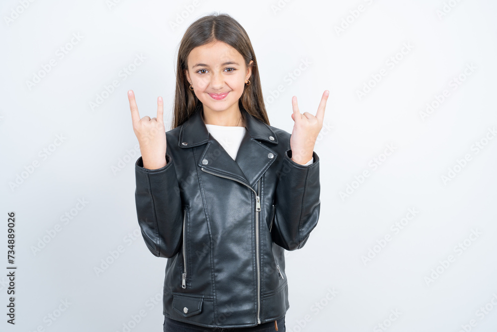 Young beautiful teen girl wearing biker jacket makes rock n roll sign looks self confident and cheerful enjoys cool music at party. Body language concept. Young beautiful teen girl wearing biker jacket makes rock n roll sign looks self confident and cheerful enjoys cool music at party. Body language concept.