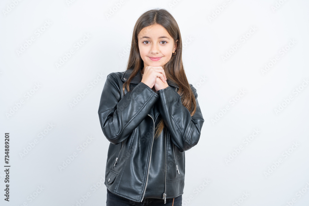 Happy Young beautiful teen girl wearing biker jacket anticipates something awesome happen, looks happily aside, keeps hands together near face, has glad expression. Happy Young beautiful teen girl wearing biker jacket anticipates something awesome happen, looks happily aside, keeps hands together near face, has glad expression.