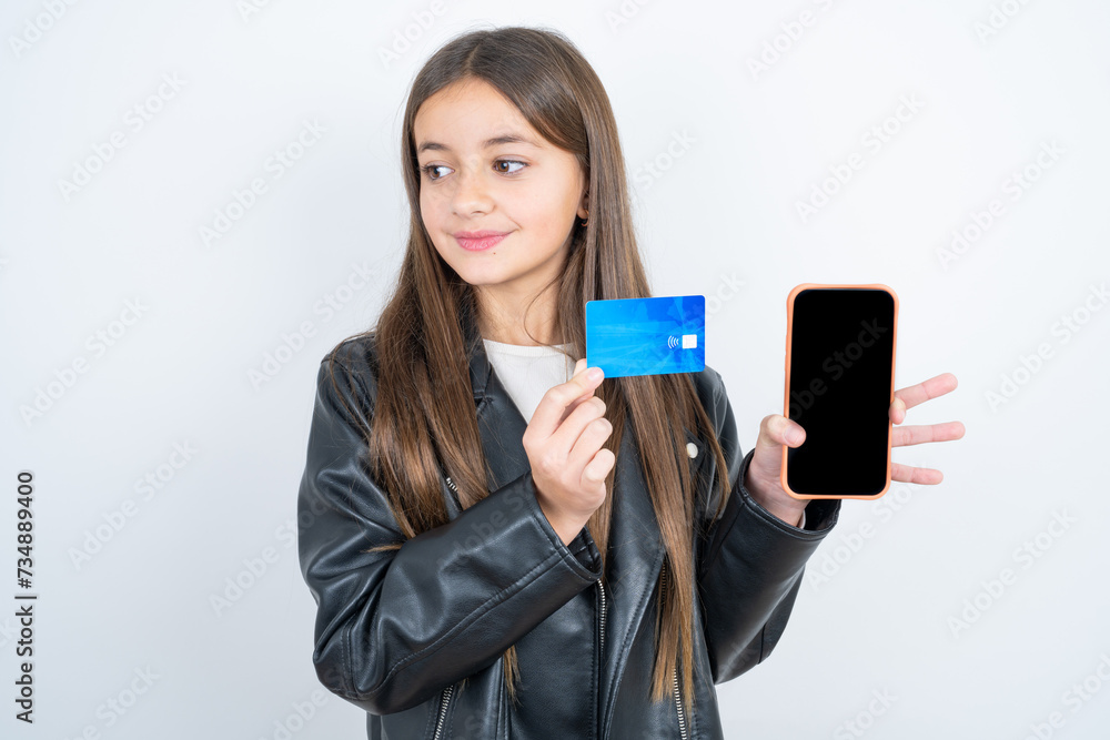 Young beautiful teen girl wearing biker jacket holding bank card modern device looking empty space Young beautiful teen girl wearing biker jacket holding bank card modern device looking empty space