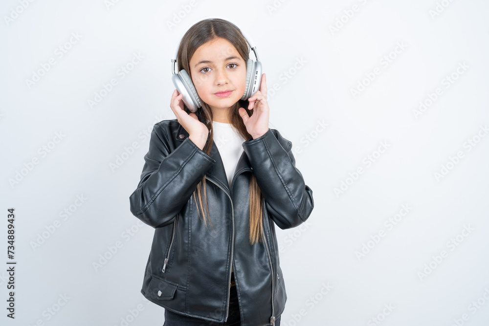 Pleased Young beautiful teen girl wearing biker jacket enjoys listening pleasant melody keeps hands on stereo headphones closes eyes. Spending free time with music Pleased Young beautiful teen girl wearing biker jacket enjoys listening pleasant melody keeps hands on stereo headphones closes eyes. Spending free time with music