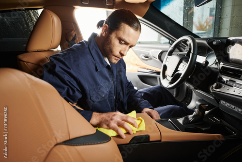 appealing devoted serviceman in blue uniform with collected hair cleaning car with yellow rag photo