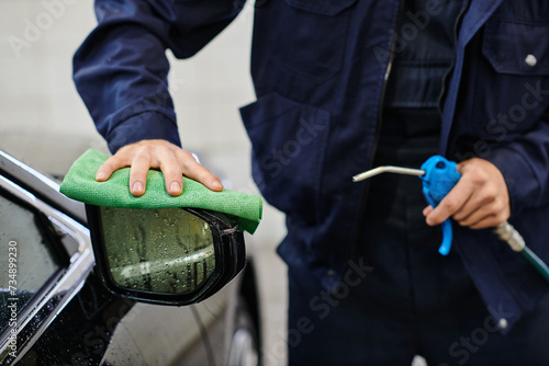 cropped view of dedicated serviceman in blue uniform cleaning car using hose and rag in garage