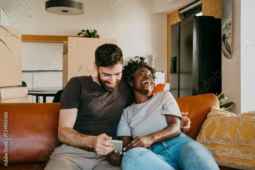 Happy multiracial couple enjoying sharing smart phone while sitting on sofa in living room at home