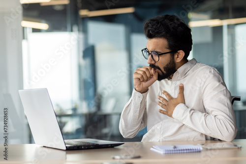 A young Indian man came down with a virus and a cold. Sitting in the office at the table and coughing, holding his chest from pain and covering his mouth with his hand