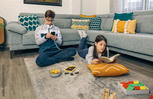 A teenage boy listens to music on his phone while his sister reads a book on the floor, surrounded by games and snacks in a cozy living room photo