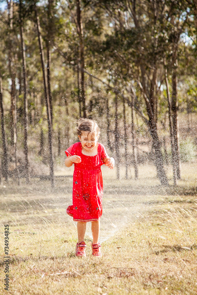 Happy little girl in pink dress laughing and running under sprinkler in backyard Happy little girl in pink dress laughing and running under sprinkler in backyard