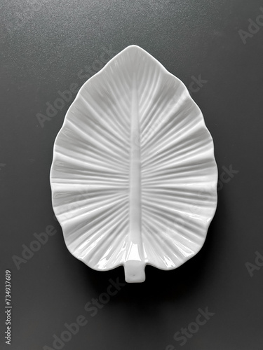 Empty white porcelain plate in shape of tree leaf and towel on dark gray background. View from above 