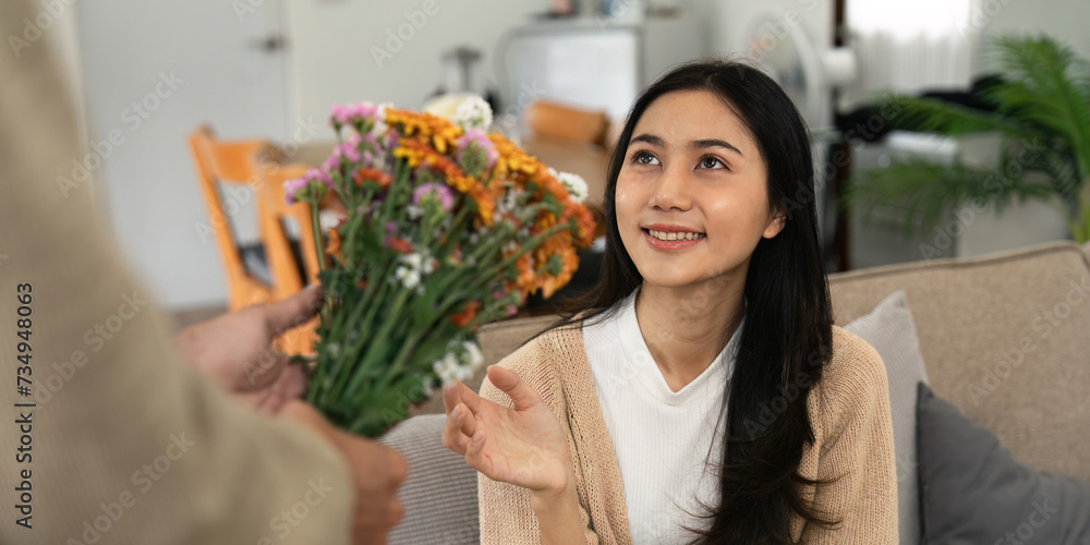 Romantic young asian couple embracing with holding flowers and smiling in living room at home. fall in love. Valentine concept