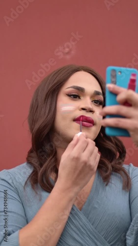 Young trans woman putting on make up and lipstick using cellphone as mirror. LGTB lifestyle concept. photo