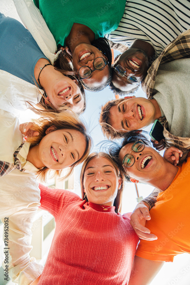 Vertical low angle view of a group of multiracial friends standing on a circle, smiling and embracing together. Young teenagers laughing and looking at camera. Team of people on a coaching meeting