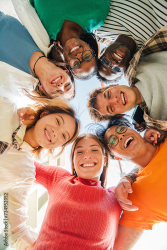 Vertical low angle view of a group of multiracial friends standing on a circle, smiling and embracing together. Young teenagers laughing and looking at camera. Team of people on a coaching meeting © Jose Calsina