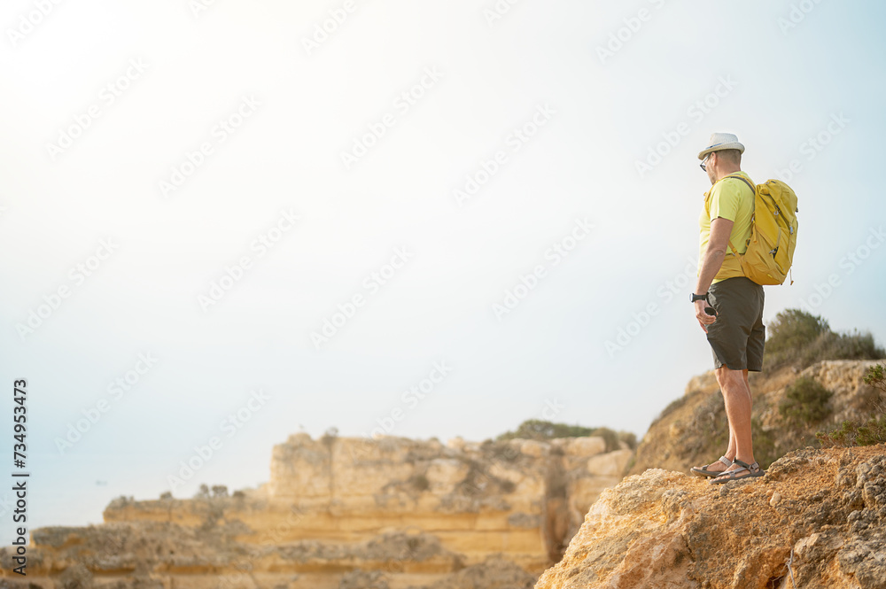 View of an unrecognizable man with backpack and hat standing ,watching the sunset from a cliff by the sea, concept of leisure and relax, copy space for text
