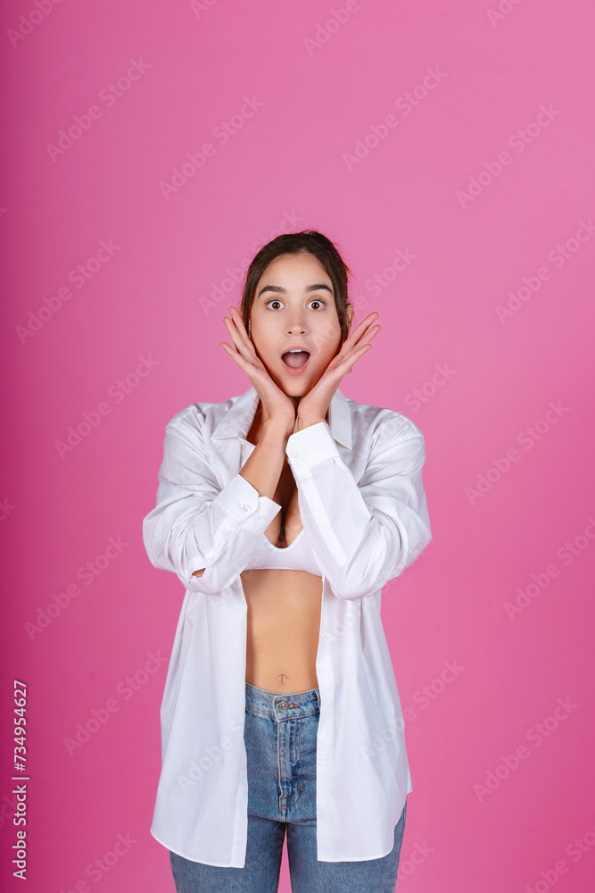 Young woman with a surprised expression, hands on cheeks, wearing a white shirt and jeans against a pink background. Young woman with a surprised expression, hands on cheeks, wearing a white shirt and jeans against a pink background.