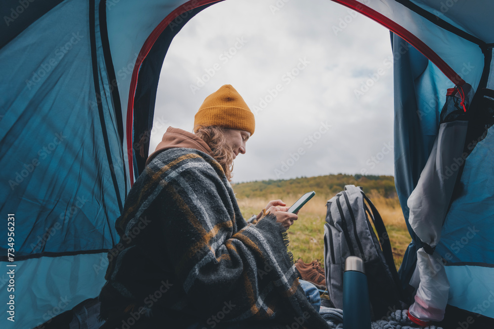 Young cheerful woman sitting in a tent and using smartphone while travelling. Hiking travel and trekking concept