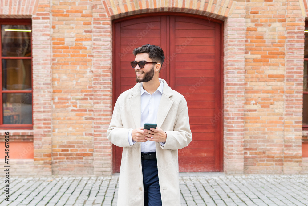 Cheerful businessman in a trench coat and sunglasses browsing on his phone, standing in front of a classic brick building with a red door Cheerful businessman in a trench coat and sunglasses browsing on his phone, standing in front of a classic brick building with a red door