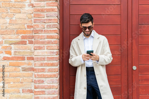 A joyful businessman in a beige coat and sunglasses using his phone, leaning against a brick wall next to a red door photo