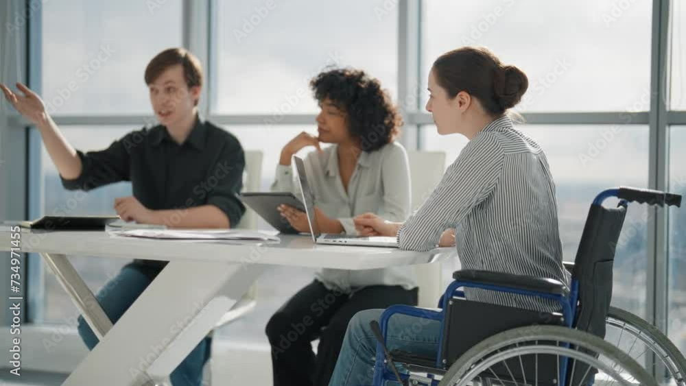 Corporate colleagues working on project in office together sitting at desk. Woman in wheelchair with physical disability on meeting with caucasian man african american female. Teamwork, discuss. Corporate colleagues working on project in office together sitting at desk. Woman in wheelchair with physical disability on meeting with caucasian man african american female. Teamwork, discuss.