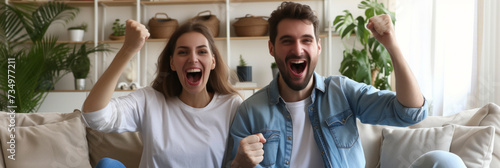 Excited African American couple celebrating success while sitting on sofa at home. Overjoyed happy young man and woman in casual wear raising clenched fists in excitement doing winner gesture photo