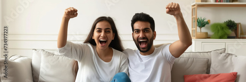 Excited Indian couple celebrating success while sitting on sofa at home. Overjoyed happy young man and woman in casual wear raising clenched fists in excitement doing winner gesture