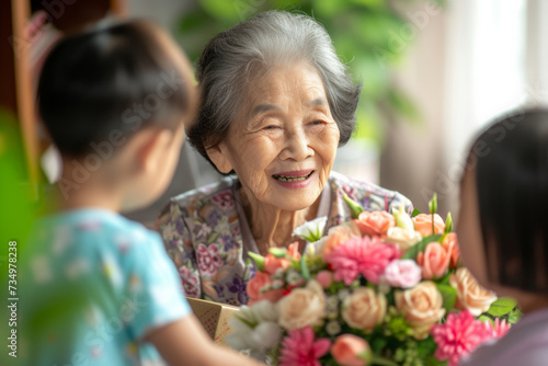 Happy senior Asian woman receives presents from her grandchildren. Children make their grandmother a birthday surprise. Little kids give their grandma a gift card and a bouquet of flowers © AI_images