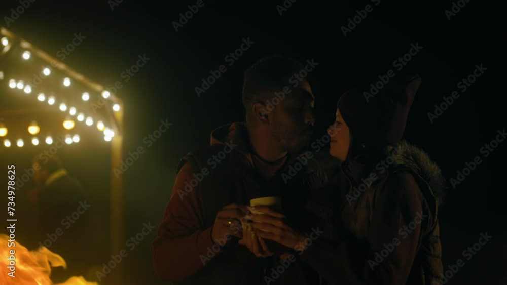 Multiethnic couple drinks tea, kisses and hugs while standing near the fire outdoors during vacation trip. In the background traveling friends rest in stylish wooden gazebo decorated with light bulbs. Multiethnic couple drinks tea, kisses and hugs while standing near the fire outdoors during vacation trip. In the background traveling friends rest in stylish wooden gazebo decorated with light bulbs.
