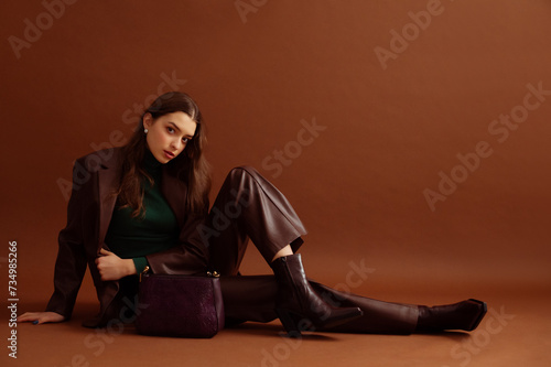 Fashionable confident woman wearing faux leather suit blazer, pants, pointed toe ankle boots, with trendy purple bag, posing on brown background. Full-length studio fashion portrait. Copy, empty space