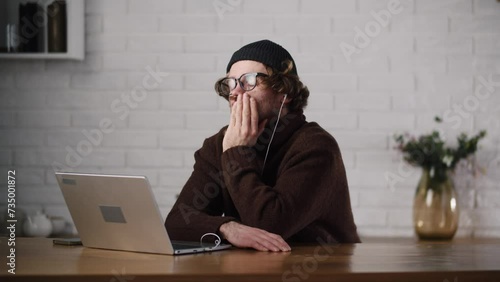 A young bored man listening lecture via laptop. Unmotivated employee feel sleepy during daily meet with team. Man listens a boring lecture at home using notebook. Uninteresting seminar online