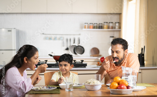 Happy parents with child eating lunch together on dining table at home - concept of weekend relaxation, family bonding and parenthood