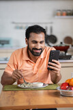 Vertical shot of Indian middle aged man busy using mobile phone while eating lunch on dining table at home - concept of modern lifestyles, internet distraction and social media sharing