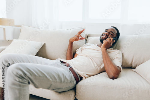 Happy African American man sitting on a black sofa, holding a smartphone and having a video call He is relaxed and confident, enjoying the convenience of modern technology at home The cozy apartment