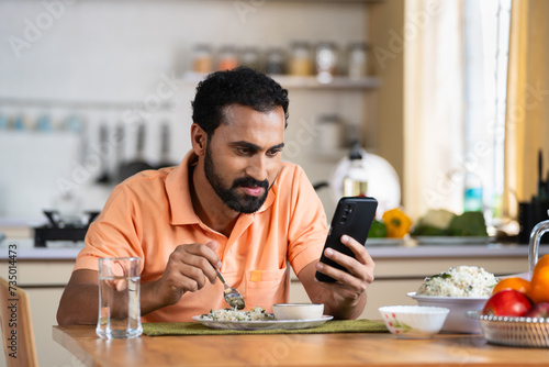 Indian middle aged man busy using mobile phone while eating lunch on dining table at home - concept of modern lifestyles, internet distraction and social media sharing