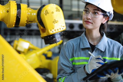 Caucasian female robotics engineer wearing helmet and safety glasses using tablet computer to inspect industrial robot arm in factory.